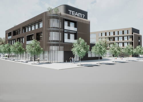 Moderner mehrstöckiger Büro- und Geschäftskomplex mit Holzlamellenfassade, großflächigen Glasfronten, TEAM7-Schriftzug und von Bäumen gesäumtem Platz mit Passanten.