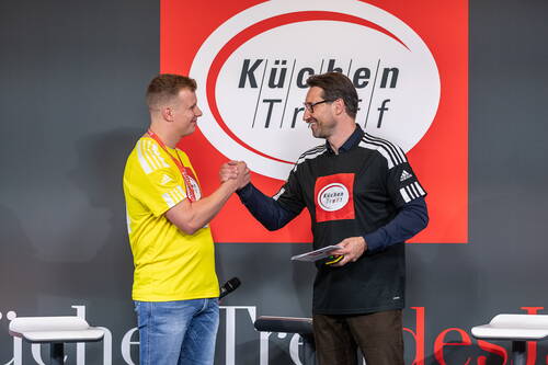Zwei Männer im KüchenTreff-Trikot schütteln sich auf einer Bühne die Hand und lächeln sich vor einem großen KüchenTreff-Logo an.