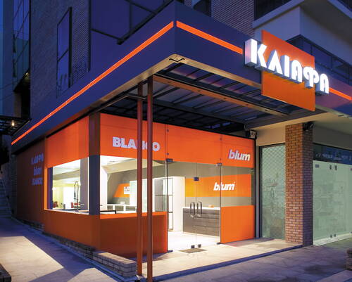 Modernes Küchenstudio mit leuchtend orange-grauer Fassadengestaltung, großen Glasfronten und beleuchteten Markenschriftzügen in einer abendlichen Straßenszene