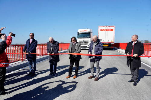 Politiker und Unternehmensvertreter durchschneiden ein rotes Band zur Einweihung einer neuen Brücke, während Lastwagen im Hintergrund auf die erste Überfahrt warten