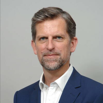 Mittelalterlicher Mann mit grauem Kurzhaar und Bart trägt dunkelblauen Anzug und weißes Hemd vor neutral grauem Hintergrund