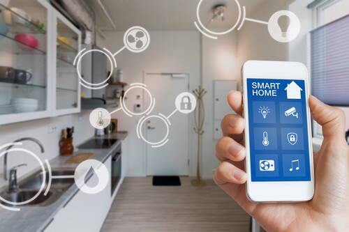 Hand hält Smartphone mit Smart‑Home‑App, die Beleuchtung, Temperatur, Sicherheit und Musik in einer modernen Küche steuert