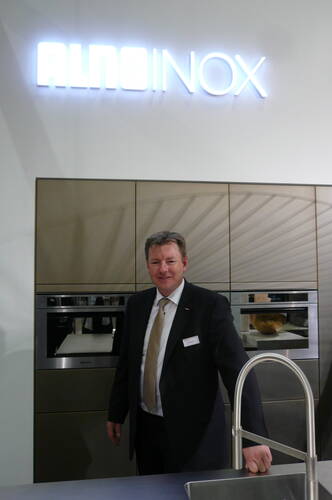 Geschäftsmann im Anzug präsentiert eine moderne Einbauküche mit Edelstahlgeräten und leuchtendem ALNO INOX Schriftzug im Hintergrund