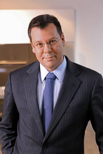 Mittelalterlicher Mann im dunklen Nadelstreifenanzug mit blauer Krawatte und Brille steht leicht lächelnd in einem modernen Bürointerieur.