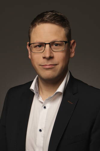 Mann mit kurzer, brauner Frisur und Brille im dunklen Anzug und weißem Hemd blickt ernst in die Kamera vor neutralem Hintergrund