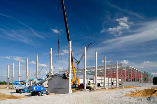 Bauarbeiter montieren mit Kränen und Hebebühnen massive Betonfertigteile am Stahlrahmen einer großen Industriehalle unter blauem Himmel