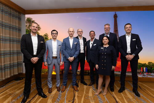 Acht geschäftlich gekleidete Personen posieren lächelnd in einem Konferenzraum vor einem großen Wandbild des Eiffelturms bei Sonnenuntergang.