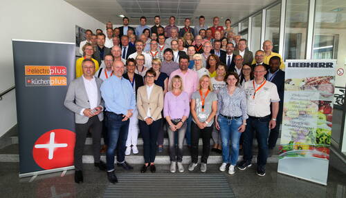 Große Gruppe von Mitarbeiterinnen und Mitarbeitern der Marken electroplus, kuechenplus und Liebherr posiert gemeinsam auf einer Treppe in einem modernen Firmengebäude während einer Veranstaltung.