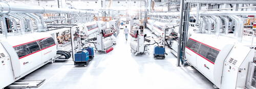 Hochautomatisierte Fertigungshalle mit parallel angeordneten Combima-II-Bearbeitungsstraßen, zahlreichen Absaugrohren und einem Techniker, der den Produktionsbereich entlanggeht