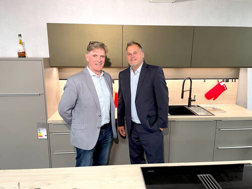 Zwei lächelnde Männer im Anzug stehen in einer modernen Einbauküche mit grauen Fronten, schwarzer Spüle und rotem Backhandschuh an der Rückwand.