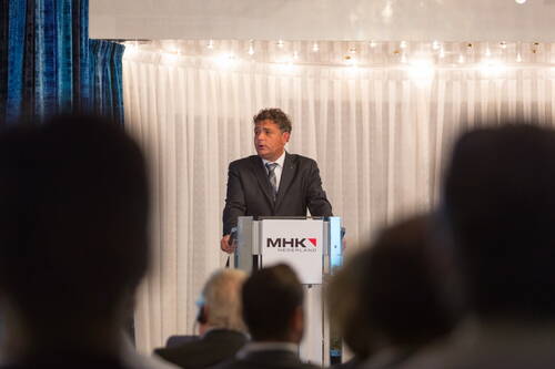 Geschäftsmann im Anzug hält eine Rede am Rednerpult von MHK Nederland vor einem sitzenden Publikum in einem hell ausgeleuchteten Konferenzsaal.