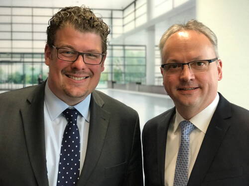 Zwei lächelnde Männer im Anzug mit Krawatte und Brille stehen nebeneinander in einer modernen, lichtdurchfluteten Bürohalle.
