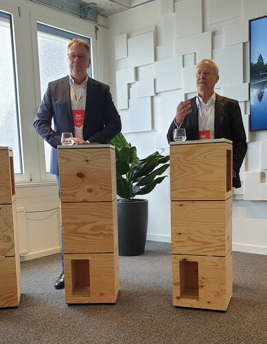 Zwei Geschäftsleute im Anzug sprechen bei einer Veranstaltung an hohen, aus Holzklötzen gebauten Rednerpulten in einem modernen Konferenzraum.