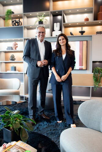 Zwei elegant gekleidete Personen stehen lächelnd in einem modern eingerichteten Büro-Loungebereich mit Designerregal, Pflanzen und gemütlichen Sitzmöbeln.