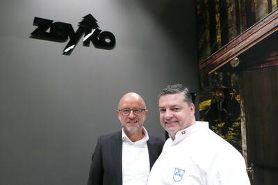 Zwei Männer lächeln in einer modernen Ausstellungshalle, links im Anzug vor einer grauen Wand mit Zeyko-Logo, rechts ein Koch in weißer Uniform mit V-Zug-Emblem vor einer Wald- und Hüttenwand.