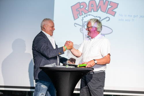 Zwei ältere Männer reichen sich auf einer Bühne lächelnd die Hände über einem Stehtisch, während im Hintergrund eine humorvolle Comic-Grafik mit dem Schriftzug „FRANZ“ projiziert wird.