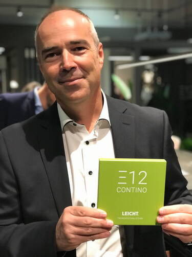 Mann im Anzug hält lächelnd ein grünes Muster mit der Aufschrift „E12 CONTINO LEICHT“ in einem modernen Ausstellungsraum in die Kamera