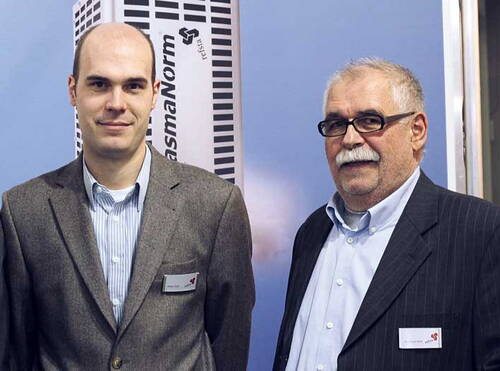 Zwei Männer im Anzug mit Namensschildern stehen nebeneinander vor einem Messestand mit der Aufschrift „PlasmaNorm“
