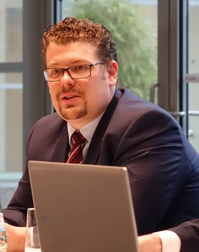 Geschäftsmann im Anzug mit roter Krawatte spricht konzentriert in einem modernen Konferenzraum hinter einem aufgeklappten Laptop