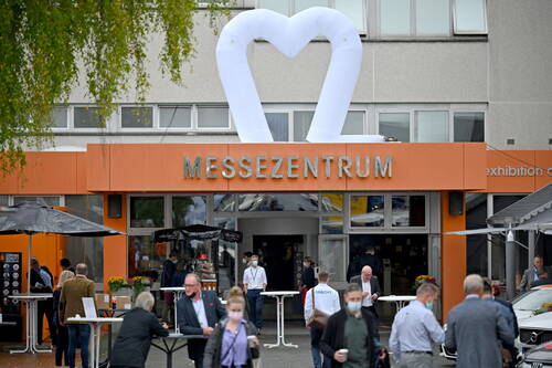 Besucher mit Mund-Nasen-Schutz strömen durch den Eingang eines orangefarbenen Messezentrums, über dem ein großes weißes aufblasbares Herz angebracht ist.