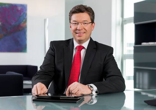 Geschäftsmann im dunklen Anzug mit roter Krawatte sitzt lächelnd an einem Glastisch in modernem Büro mit abstraktem Wandbild im Hintergrund