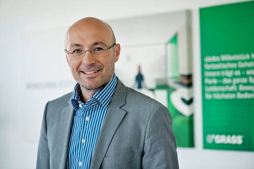 Geschäftsmann mit Brille und grauem Anzug lächelt in modernem Büro mit grünen Firmenpostern im Hintergrund