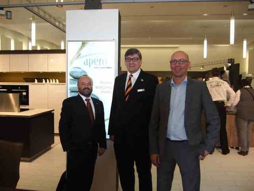 Drei Männer in Anzugkleidung posieren lächelnd in einer modernen Ausstellungshalle vor einem beleuchteten Werbedisplay und einer offenen Küchenzeile.