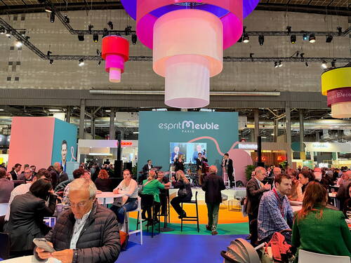 Menschenmenge auf der Möbelmesse „Esprit Meuble“ in Paris, die an Stehtischen sitzt, sich unterhält und einem Bühnenprogramm unter bunten, von der Decke hängenden Leuchtelementen zuschaut