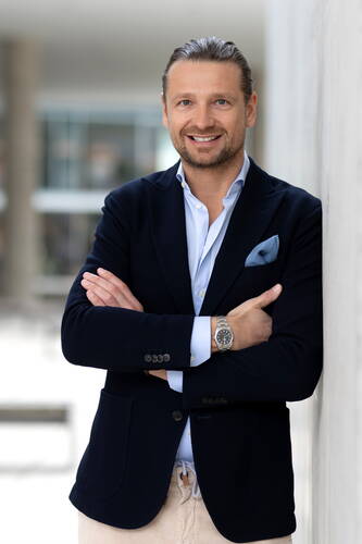 Lächelnder Mann mit zurückgekämmten Haaren, im dunkelblauen Sakko und hellblauen Hemd, steht mit verschränkten Armen an einer hellen Wand in moderner Büroumgebung