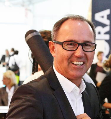 Lächelnder Mann im Anzug mit Brille trägt eine schwarze Posterrolle über der Schulter in einer geschäftigen Messehalle