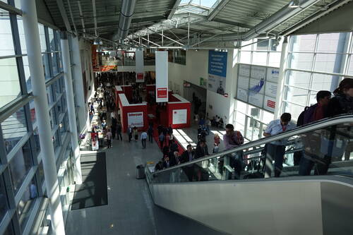 Besucher strömen durch die helle Messehalle mit roten Interzum-Ständen, während Menschen auf einer Rolltreppe in den Ausstellungsbereich hinabfahren.