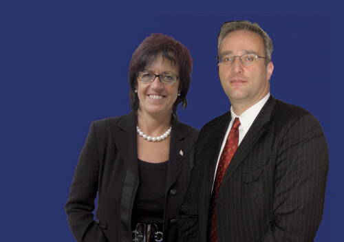 Zwei geschäftlich gekleidete Personen mit Brille stehen vor einfarbig blauem Hintergrund und blicken in die Kamera