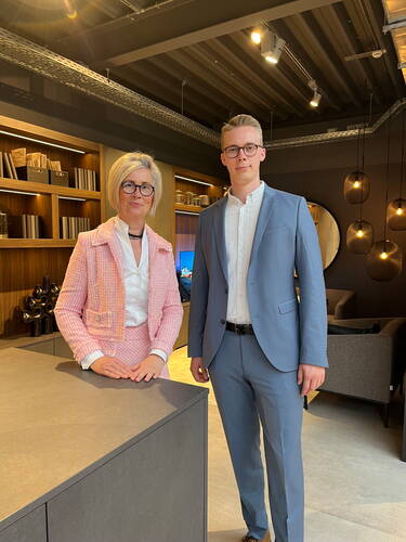 Zwei elegant gekleidete Personen in Anzügen stehen in einem modern eingerichteten Bürobereich und blicken direkt in die Kamera.