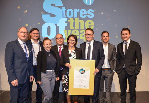 Gruppe von Geschäftsleuten im Anzug posiert auf einer Bühne bei der Preisverleihung „Stores of the Year“, während zwei Personen eine große gerahmte Urkunde in der Mitte halten