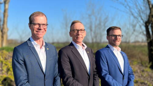 Drei lächelnde Männer in Anzügen mit Brille posieren nebeneinander im Freien vor unscharfer Landschaft bei blauem Himmel