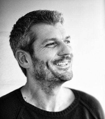 Lächelnder Mann mittleren Alters mit Bart und kurzen Haaren blickt seitlich nach rechts in einem schwarz‑weißen Porträt