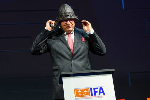 Redner im Anzug setzt sich auf der IFA-Bühne eine lederne Pilotenhaube auf und steht hinter einem Rednerpult mit IFA-Logo