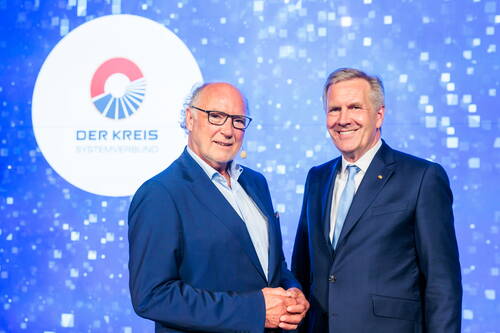 Zwei elegant gekleidete Männer in blauen Anzügen lächeln vor einer Bühne mit dem Logo „DER KREIS Systemverbund“ auf blauem, pixelartigem Hintergrund