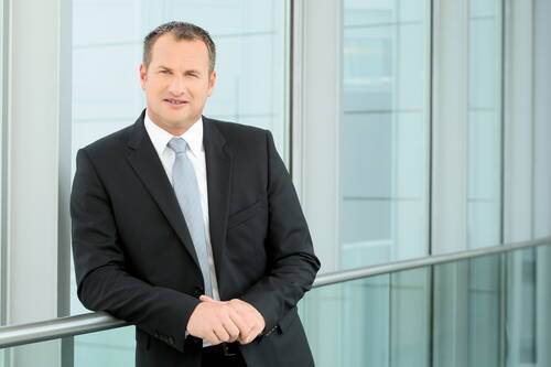 Geschäftsmann im schwarzen Anzug lehnt entspannt am Glasgeländer eines modernen Bürogebäudes und blickt selbstbewusst in die Kamera