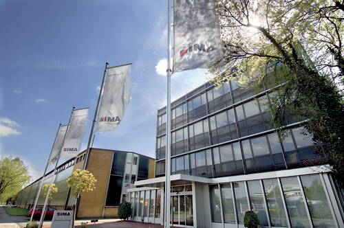Moderne Firmenzentrale von IMA mit verglaster Fassade und mehreren wehenden Unternehmensflaggen an einem sonnigen Frühlingstag