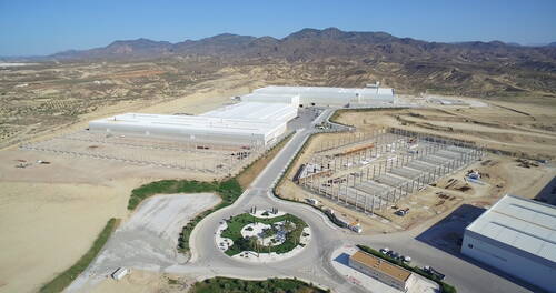Großes Industrie- und Logistikgelände in karger Berglandschaft mit mehreren weißen Hallen, einem begrünten Kreisverkehr im Vordergrund und einer sich im Bau befindlichen Stahlkonstruktion.