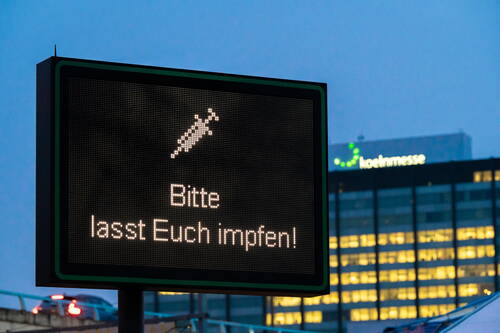 Digitale Anzeigetafel in der Dämmerung mit der deutschsprachigen Aufforderung „Bitte lasst Euch impfen!“ vor beleuchtetem Bürogebäude