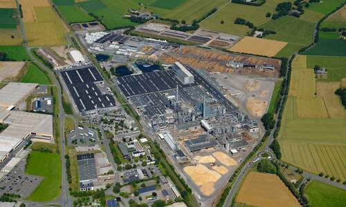 Großes Industrie- und Sägewerksgelände mit Lagerflächen für Holzspäne und Rundholz, umgeben von grünen Feldern und Zufahrtsstraßen aus der Luftperspektive