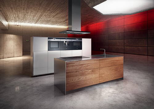 Minimalistische Designküche mit freistehender Kochinsel aus Holz, integrierter Induktionskochfläche, Deckenhaube und weißem Geräteblock vor rot beleuchteter Betonwand