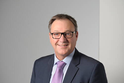 Älterer Mann mit Brille im dunklen Anzug und rosa Krawatte lächelt vor neutral grauem Hintergrund in die Kamera