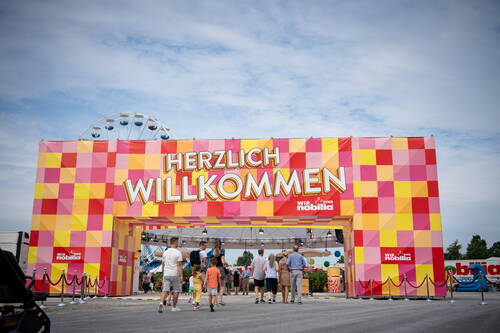 Bunte Eingangspforte mit der Aufschrift „Herzlich Willkommen“, durch die Familien auf ein belebtes Festgelände mit Riesenrad im Hintergrund gehen.