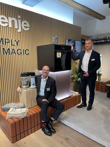 Zwei Männer im Anzug präsentieren in einer modern gestalteten Ausstellungsküche einen schwarzen Einbauofen und eine Küchenmaschine auf einem Podest vor einer Holzlamellenwand mit Gorenje-Logo.