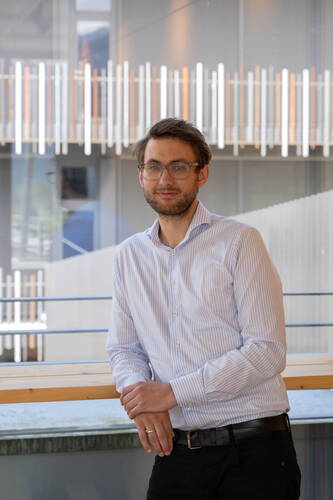 Junger Mann mit Brille und gestreiftem Hemd lehnt lässig an einem Holztisch vor einer modernen Glasfassade mit vertikalen Lichtleisten im Hintergrund