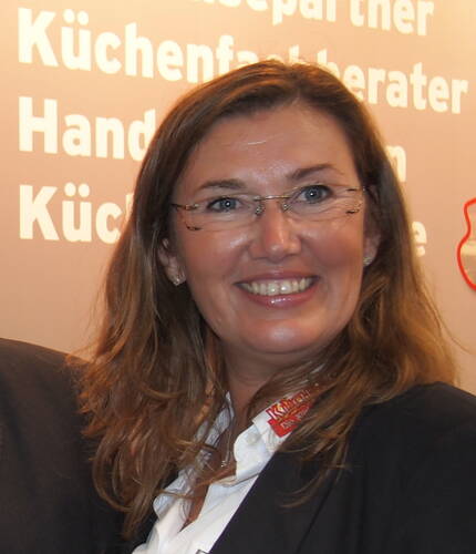 Lächelnde Frau mit Brille und langen braunen Haaren in Businesskleidung vor einer beschrifteten Messewand