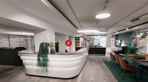 Modern gestalteter Empfangsbereich mit geschwungener weißer Theke, dekorativen Pflanzen, stilvollen Deckenleuchten und angrenzender Sitz- und Beratungsecke in einem Büro- oder Showroom.